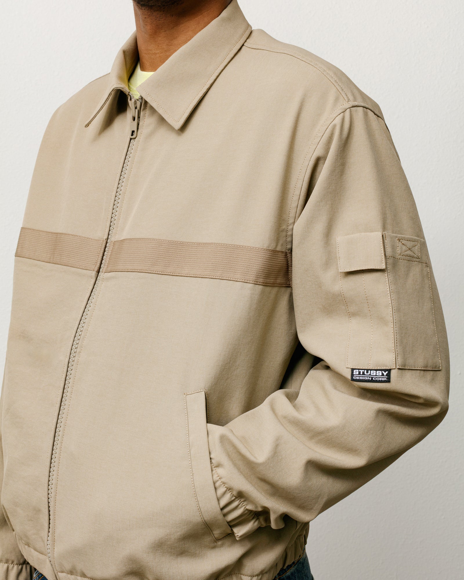 Khaki mechanic jacket with zip front, webbing stripe, and a sleeve patch pocket.