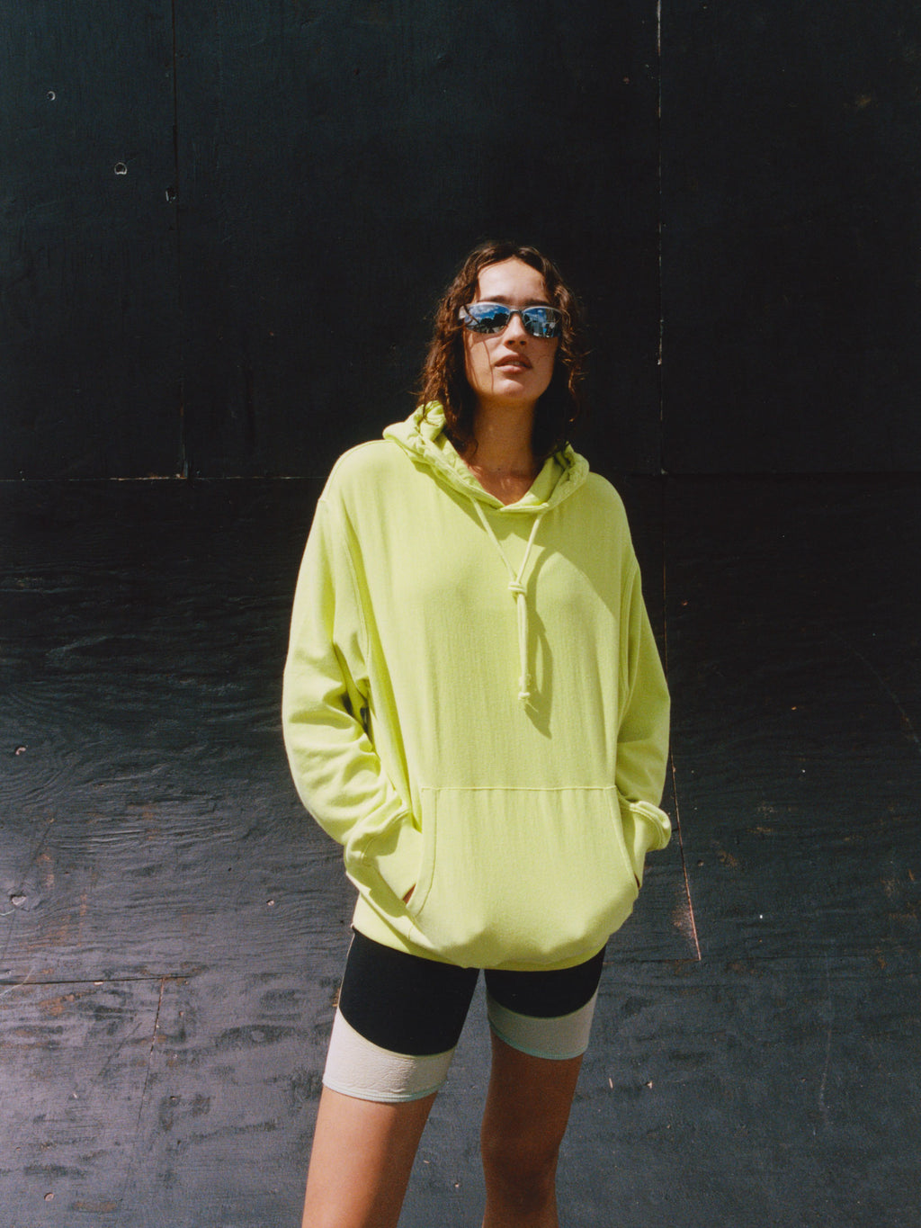 Model wearing the Lightweight Terry Hoodie in Yellow with bike shorts and sunglasses against a dark wooden wall.