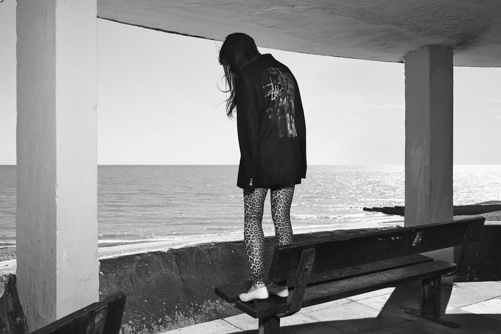A black-and-white photo showing the back of a person standing on a bench overlooking the sea, wearing the JUNYA SPORT COAT and leopard-print tights.